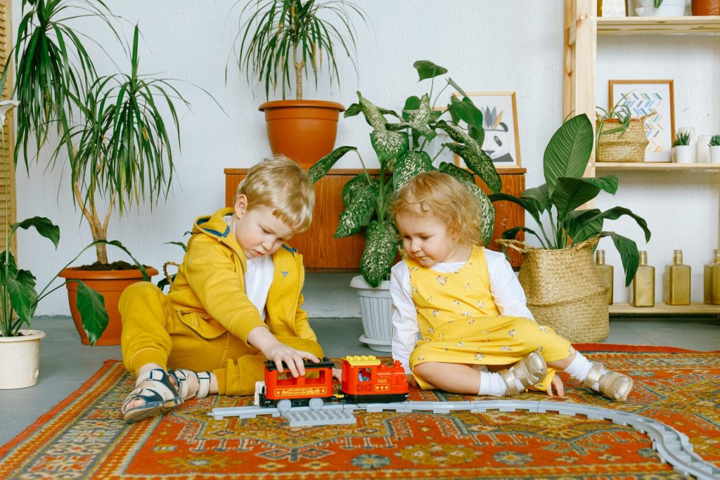 Two children enjoy playing with a toy train on a colorful rug indoors surrounded by plants.